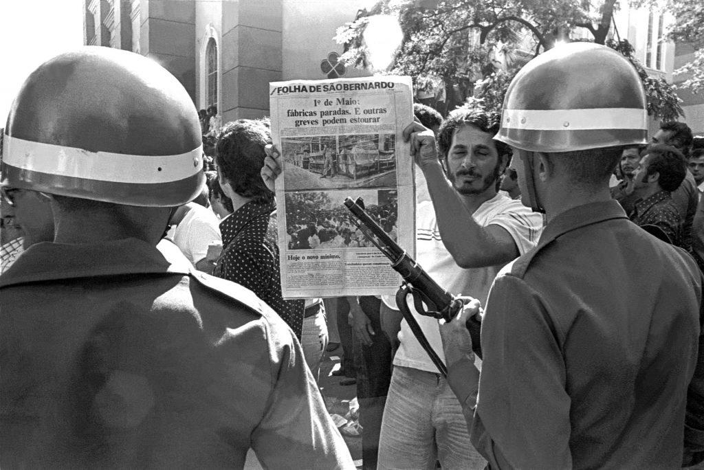 Durante a manifestação do 1º de Maio de 1980, um metalurgista em greve confronta soldados exibindo manchete sobre fábricas paradas. A cena expressa a resistência operária frente à repressão apoiada por empresas que financiaram e colaboraram com o regime militar para reprimir greves e movimentos sindicais.
