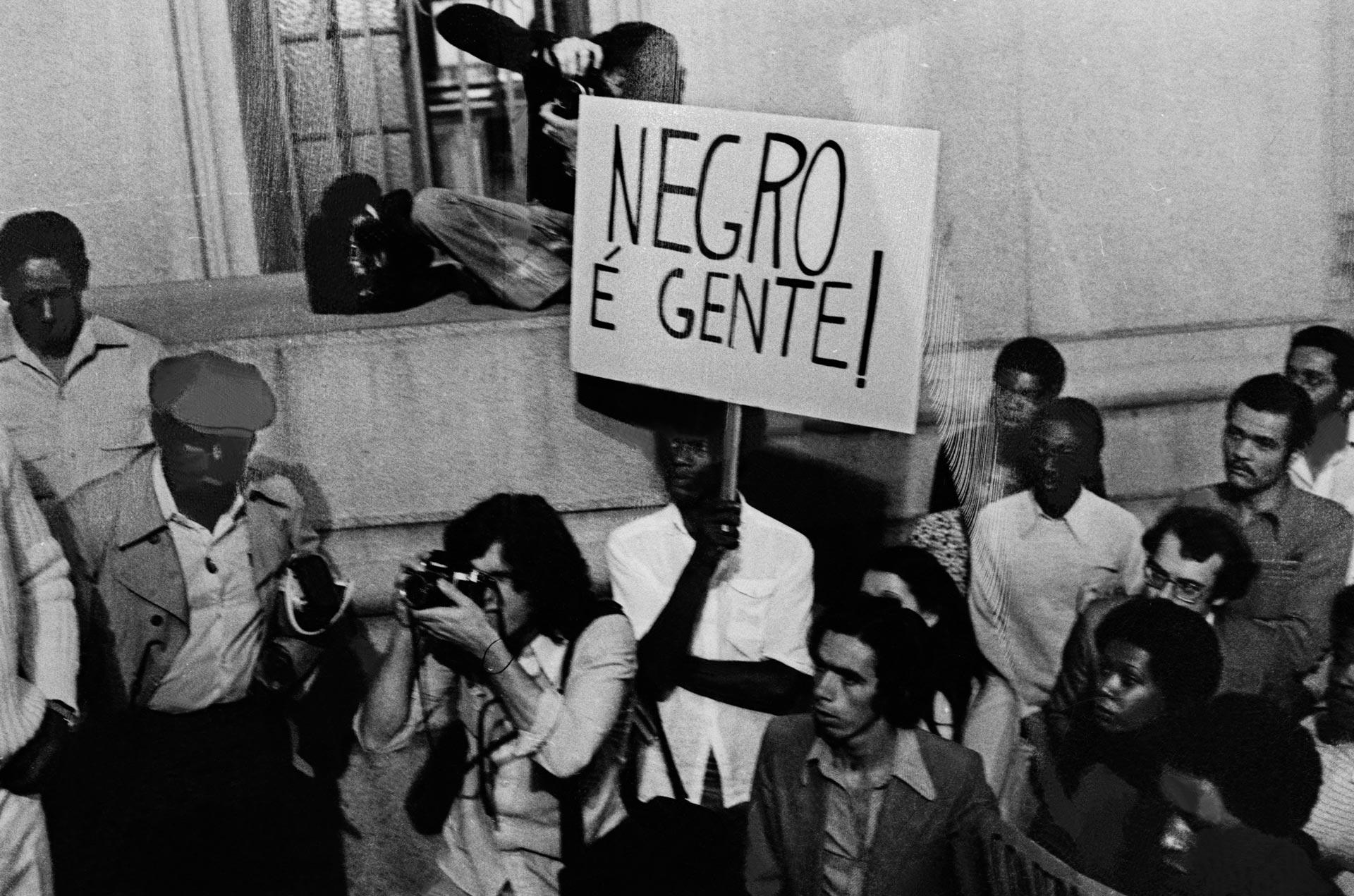 Manifestante do Movimento Negro Unificado (MNU) em ato no centro de São Paulo, em 1980. O cartaz com a frase "Negro é Gente!" sintetiza a luta do movimento contra a violência e a desumanização da população negra, denunciando a falácia da "democracia racial" durante a Ditadura Militar.